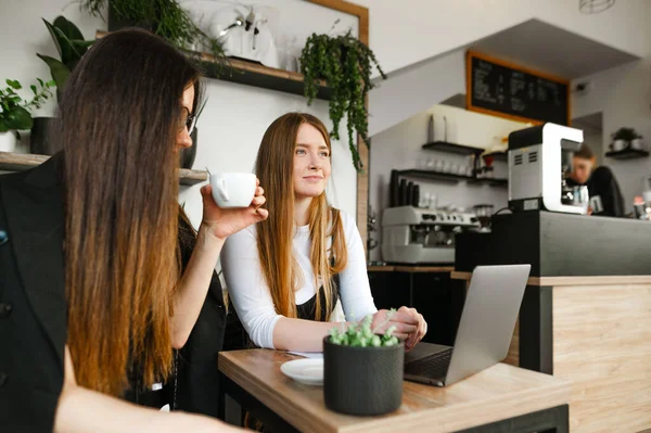 Resmi kıyafetli iki kız arkadaş parlak bir hipster kafede dizüstü bilgisayarlarıyla oturup kahve içip sohbet ediyorlar. İki ofis çalışanı bir kafede dinleniyorlar. İş toplantısı.