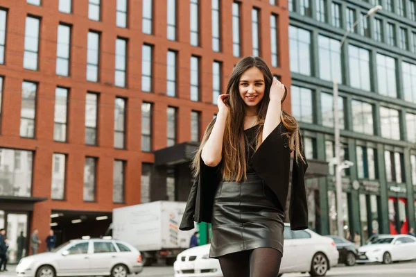 Happy young business woman in dark formal wear standing on the street and posing at camera on cityscape background.Smiling lady in casual clothes stands on the street,background of modern architecture