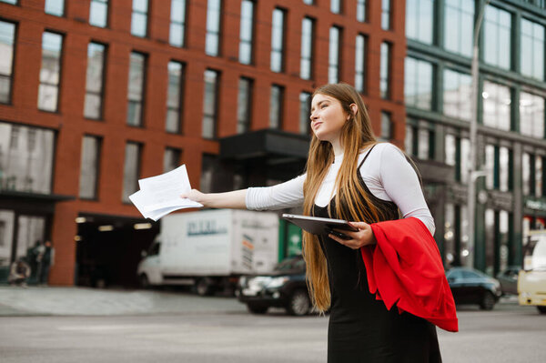 Busy female student stands with a laptop and papers in her hands and catches a taxi hand gesture with a smile on her face. Positive girl in formal clothes stops a taxi on the street of a metropolis.