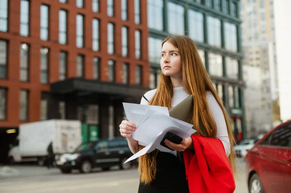 Portrait of a cute student girl standing on the street of a big city with laptop, smartphone, notes in hands,looking away with a serious face.Student walking down the street with papers and gadgets