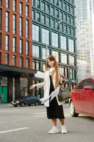 Student girl in casual clothes stands on the street with a bag in her hands and misses the paper in astonishment. Lady with long hair throws paper down on cityscape background.