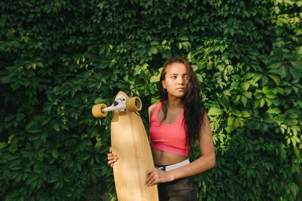 Attractive girl in casual sportswear stands on a background of green ivy with a longboard in her hand and looks away with a serious face. Fashionable hispanic girl holding a longboard in her hands