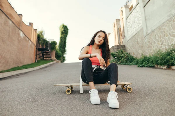カジュアルな服を着たスタイリッシュな女の子の肖像画は 通りのスケートに座って カメラを見ています 道路上のログボードに座ってリラックスした魅力的なヒスパニック系の女の子 ストリートスタイル — ストック写真