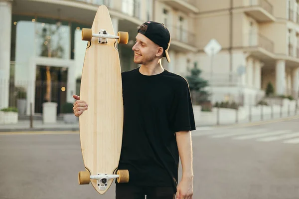 Positive guy in black clothes stands with a longboard in his hands on background of the street of the town, looks at the board with a smile on his face.Cheerful skater happy with longboard. Skating