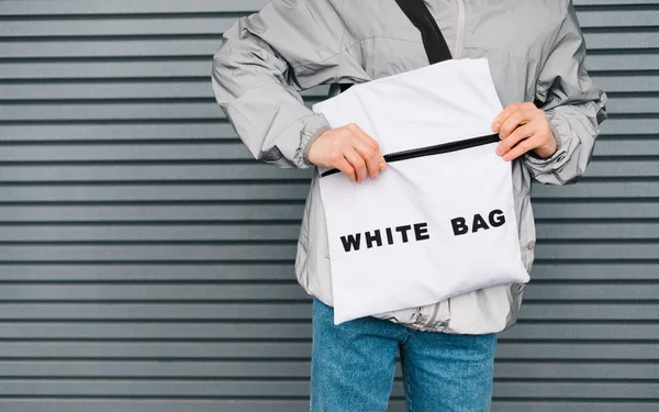 Closeup photo of a white eco bag in the hands of a young stylish man on a gray wall background, with his hand opens the zipper lock in eco bag. Reusable stylish shopping bag in the hands of a man.