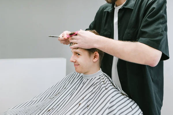 幸せな若いクライアントは 男性の美容室で椅子に座って 理容室で髪をカットします プロの男性の美容師は白い壁の背景に肯定的な若い男をカットします スペースのコピー — ストック写真