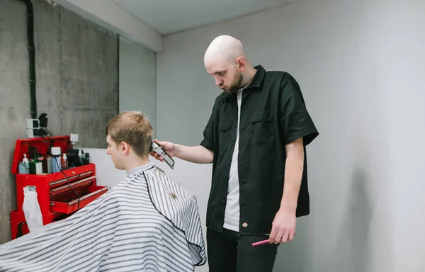 Bald man hairdresser in a black shirt cuts the hair of a young man with a clipper in his hands. Professional male hairdresser makes a stylish haircut for a barber shop with gray walls.