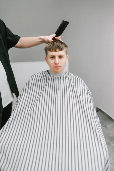 Młody Klient Zakładu Fryzjerskiego Siedzący Krześle Peignir Szarym Tle Patrzący — Zdjęcie stockowe