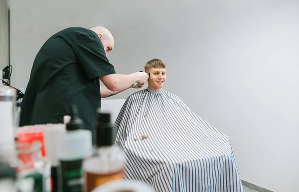 Smiling young man sitting in hairdresser chair and cutting hair at hairdresser, barber uses hair clipper, on gray background.Process of creating a stylish hairstyle in a men\'s shop,client is satisfied