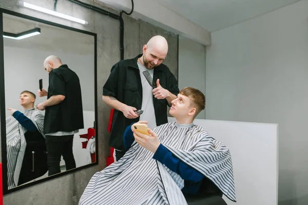 Positive young man sitting in hairdresser chair and showing barber references hairstyles on smartphone, hairdresser showing thumb up. Smiling barber makes a stylish hairstyle for a guy.