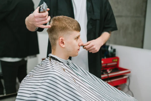 专业理发师剪头发的年轻金发男子的假发 客户坐在理发店与平静的脸 并看向别处 穿着黑色衬衫的男性理发师为男人创造时尚的发型 — 图库照片