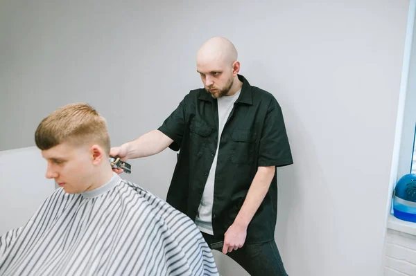 真剣なプロのヘアカッターは 彼の手にクリップで若い男を切断し 光の壁に対して彼の頭を熱心に見ています 貧しい理髪店は若い顧客を削減します スペースのコピー — ストック写真