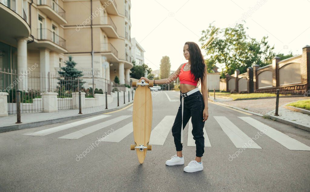 Street girl in stylish summer clothes stands on the street with a longboard in her hands on a sunset background, looking away. Attractive latin girl posing on camera with longbord in her hands.