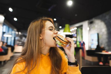 Gözlüklü ve sarı saçlı güzel bir kızın yakın plan portresi bir fast food restoranında iştah açıcı bir burgeri ısırır ve tatmin olur. Kafede abur cubur yiyen kız..