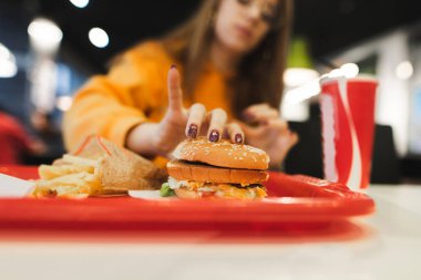 Yakın çekim. Bir kızın eli tepsiden bir hamburger alır. Günlük giysiler içindeki kadın kafede abur cubur yiyor. Mola için fast food yiyen, patates kızartması yiyen, sandviç yiyen ve kola içen bir kız.