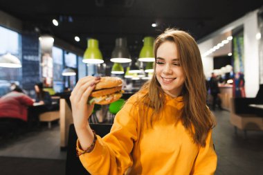 Fast food restoranında elinde hamburgerle oturmuş lezzetli abur cuburlara bakıp gülümseyen mutlu bir kızın portresi. Öğrenci kız kafede burger yiyor..