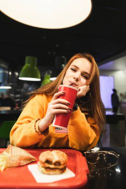 Fast food restoranında abur cubur yiyen, kameraya bakıp kırmızı bardaktan soda içen çekici bir kadının portresi. Kafede fast food yiyip kameraya poz veren tatlı bir kız..