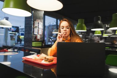 Güzel kız öğrenci kafede fast food yiyor, dizüstü bilgisayar kullanıyor, patates kızartması yiyor ve ekrana bakıyor. Şirin bayan fast food restoranındaki bir bilgisayarda çalışıyor ve abur cubur yiyor..