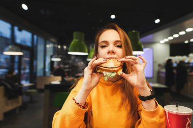 Turuncu kazaklı çekici bir kız kafede hamburger yer, kameraya bakar. Fast food restoranında abur cubur yiyen güzel kız elinde çizburger tutuyor..
