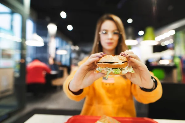 女孩坐在咖啡店 手里拿着汉堡 在咖啡店吃午饭的漂亮姑娘手里拿着汉堡包的合影 复制空间 — 图库照片