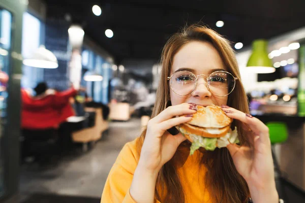 眼鏡とブロンドの髪のきれいな女の子は 口の中にハンバーガーを食べ 深刻な顔でカメラを見て レディーは ファーストフードレストランの背景に大きなハンバーガーをかむ ファーストフードランチ — ストック写真