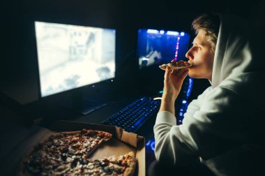 Gündelik giysiler içindeki aç oyuncu geceleri bilgisayarının başına oturur, oyunlar oynar ve kutudan pizza yer. Pizza yiyip bilgisayar kullanan genç bir adamın portresi. Oyun ve fast food..