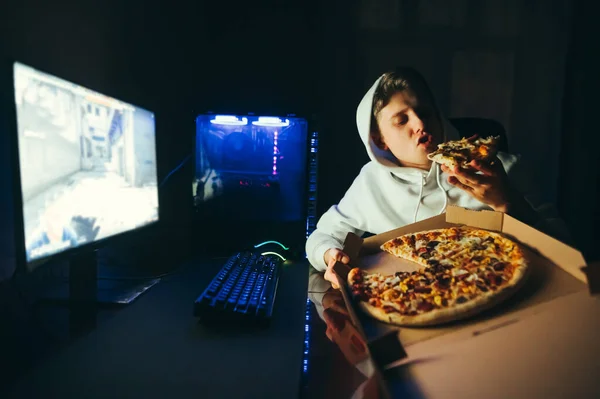 Casual gamer sits at a table at home near a computer and eats delicious pizza out of the box. Hungry young man relaxing sitting at his computer, eating pizza at home at night. Fast food and gaming.