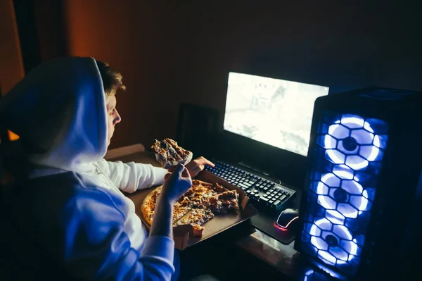 Man in hoodie sits at a table at night, plays video games on a computer and eats pizza from a box, holds a piece in his hand. Gamer and computer games at night with pizza. A hungry guy plays games