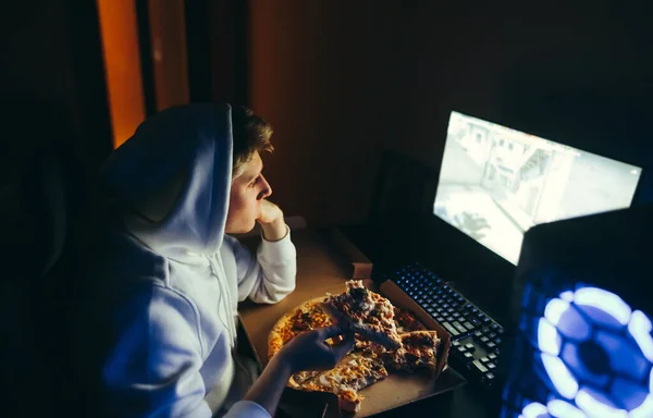 Young man sits at night at a table, watches a video on a computer and eats pizza from a hoarder. Guy teenager in a hoodie booze having pizza at the computer. Fast food and gamer.