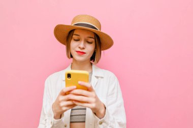 Pembe arka planda izole edilmiş, güneş şapkalı ve açık renk elbiseli bir bayan ciddi görünümlü bir akıllı telefon kullanıyor. Şirin bayan interneti akıllı telefonunda kullanıyor..