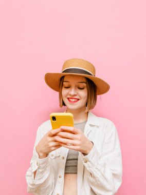 Şapkalı mutlu bayan akıllı telefon kullanıyor ve yüzünde bir gülümsemeyle ekrana bakıyor. Yüzünde gülümseme olan akıllı telefon kullanan bir tatilci kızın dikey portresi. İzole edilmiş.