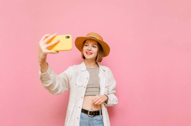 Yüzünde bir gülümseme ve kafasında güneş şapkası olan sevimli bir kız, yazlık kıyafetler giyiyor, pembe arka planda özçekim yapıyor, mutlu bir yüzle kameraya bakıyor. Yakın plan kadın selfiea alıyor.