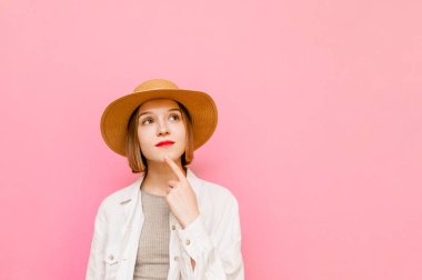 Şapkalı ve açık renk elbiseli kız pembe arka planda izole edilmiş, başka tarafa bakıyor ve düşünüyor. Pastel pembe arka planda, yaz kıyafetleri giymiş düşünceli bir kadının portresi. Boşluğu kopyala