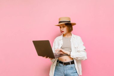 Şapkalı ve yaz elbiseli konsantre kız pembe arka planda dizüstü bilgisayar kullanıyor. Laptopta çalışan çekici bayan turist pembe arka planda izole edilmiş. Boşluğu kopyala