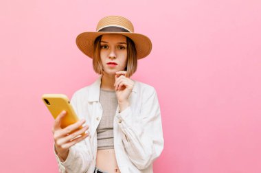 Ciddi bir yüzü olan güzel bir kız elinde akıllı telefonuyla pembe arka planda duruyor, ciddi bir yüzle kameraya bakıyor, şapka takıyor ve yaz kıyafetleri giyiyor. Boşluğu kopyala