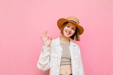 Şapkalı ve açık renk elbiseli güzel bir kız jest yapıyor ve pembe arka plan ve göz kırparak kameraya bakıyor. Çekici bayan turist yüzünde bir gülümsemeyle iyi bir jest gösteriyor. Dikey.