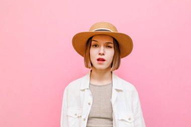 Pembe arka planda komik bir kızın yakın plan portresi, şaşırmış bir suratla kameraya bakıyor ve gözlerini kapatıyor, şapka takıyor ve yaz kıyafetleri giyiyor. İzole edilmiş.