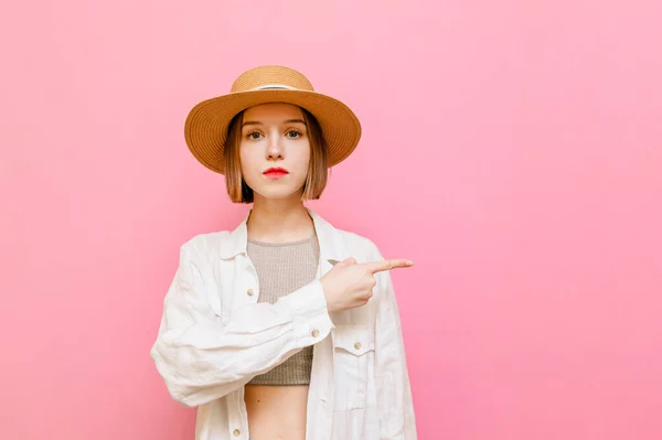 Serious lady in a hat and light summer clothes, looks into the camera and points her finger to the side. Concentrated girl shows aside on copy space isolated on pink background.