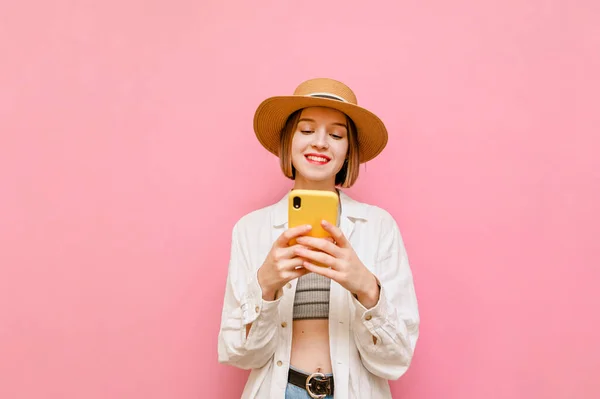 明るい服を着た幸せな女の子とピンクの背景に立つ太陽の帽子は 彼女の顔に笑顔でスマートフォンを使用しています 女性は電話や笑顔でインターネットを利用しています — ストック写真