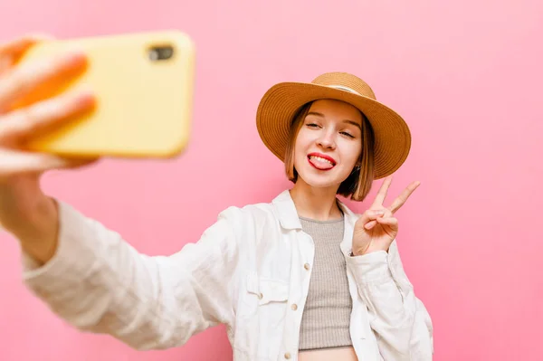 帽子と夏の光の服の幸せな女性は スマートフォンのカメラで写真を撮り 笑顔とピンクの背景に隔離された平和のジェスチャーを示しています 可愛いです笑顔女の子取りますA自撮り上のパステルの背景 — ストック写真