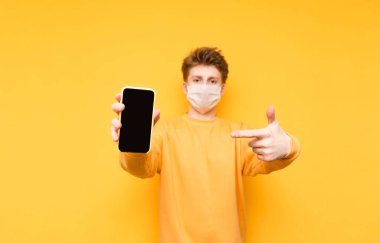 Sarı arka planda izole edilmiş tıbbi koruyucu maskeli genç bir adam kameraya bakıyor, siyah ekranlı bir akıllı telefon tutuyor ve parmağını işaret ediyor. Koronavirüs salgını. Karantinaya alın. covid-19.