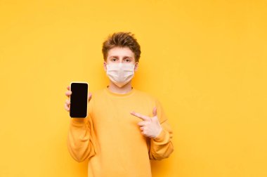 Gazlı bez maskeli yakışıklı bir adam sarı arka planda elinde akıllı bir telefonla duruyor, parmağını siyah ekrana doğrultuyor ve ciddi bir yüzle kameraya bakıyor..