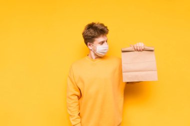 Günlük giysiler içinde ve tıbbi maske takmış yakışıklı bir genç elinde kese kağıdıyla sarı bir arka planda duruyor. Yemek teslimatı konsepti. Koronavirüs salgını. Karantinaya alın. Covid- c.