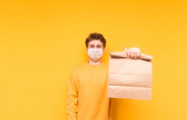 Koruyucu tıbbi maskeli yakışıklı bir genç sarı arka planda duruyor teslimattan bir paket yiyecekle, kameraya bakıyor ve gülümsüyor. Karantinaya alındı.