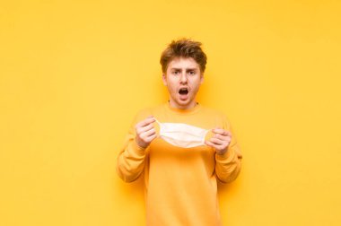 Elinde gazlı bez maskesiyle şok olmuş genç bir adam sarı arka planda izole edilmiş, şaşırmış bir yüzle kameraya bakıyor. Koronavirüs salgını. Karantinaya alın. covid-19.