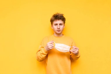 Sarı kazaklı yakışıklı genç adam elinde tıbbi gazlı bez maskesiyle sarı arka planda duruyor ve ciddi bir yüzle kameraya bakıyor. Adam sarı arka planda gazlı bez maskesi takıyor..