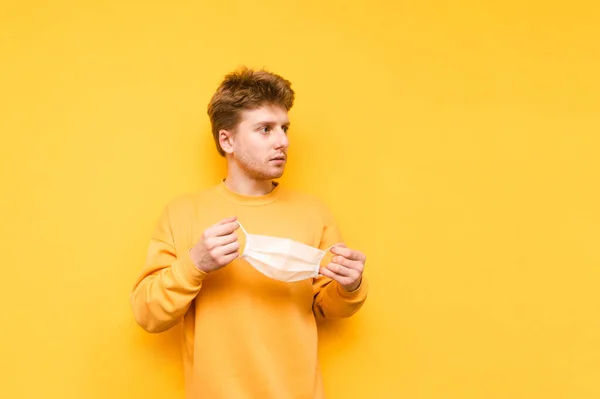 Young man in casual clothes stands on a yellow background with a medical mask in his hands and looks away with a serious face.Guy took off a protective gauze mask.Coronavirus pandemic. Quarantine