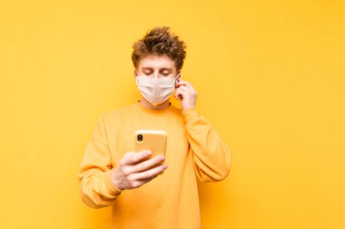 Gazlı bez maskeli genç bir adam ve... Kablosuz kulaklık sarı arka planda müzik dinliyor ve akıllı telefon kullanıyor. Koronavirüs salgını. COVID-19. Karantina.