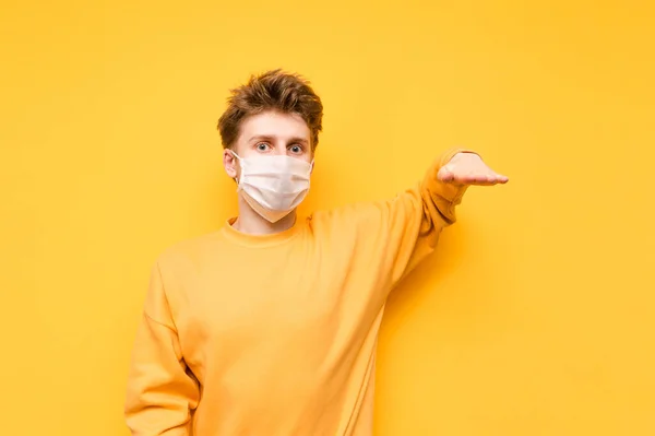 stock image Guy in a medical gauze mask holds a copy space with his hand and looks at the camera with a shocked face, isolated on a yellow background. Coronavirus pandemic. COVID-19. Quarantine.