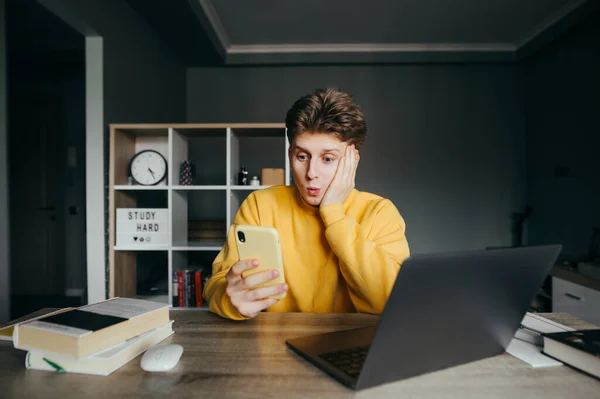 Surprised Young Man Sitting Desktop Home Laptop Books Uses Smartphone — Stock Photo, Image